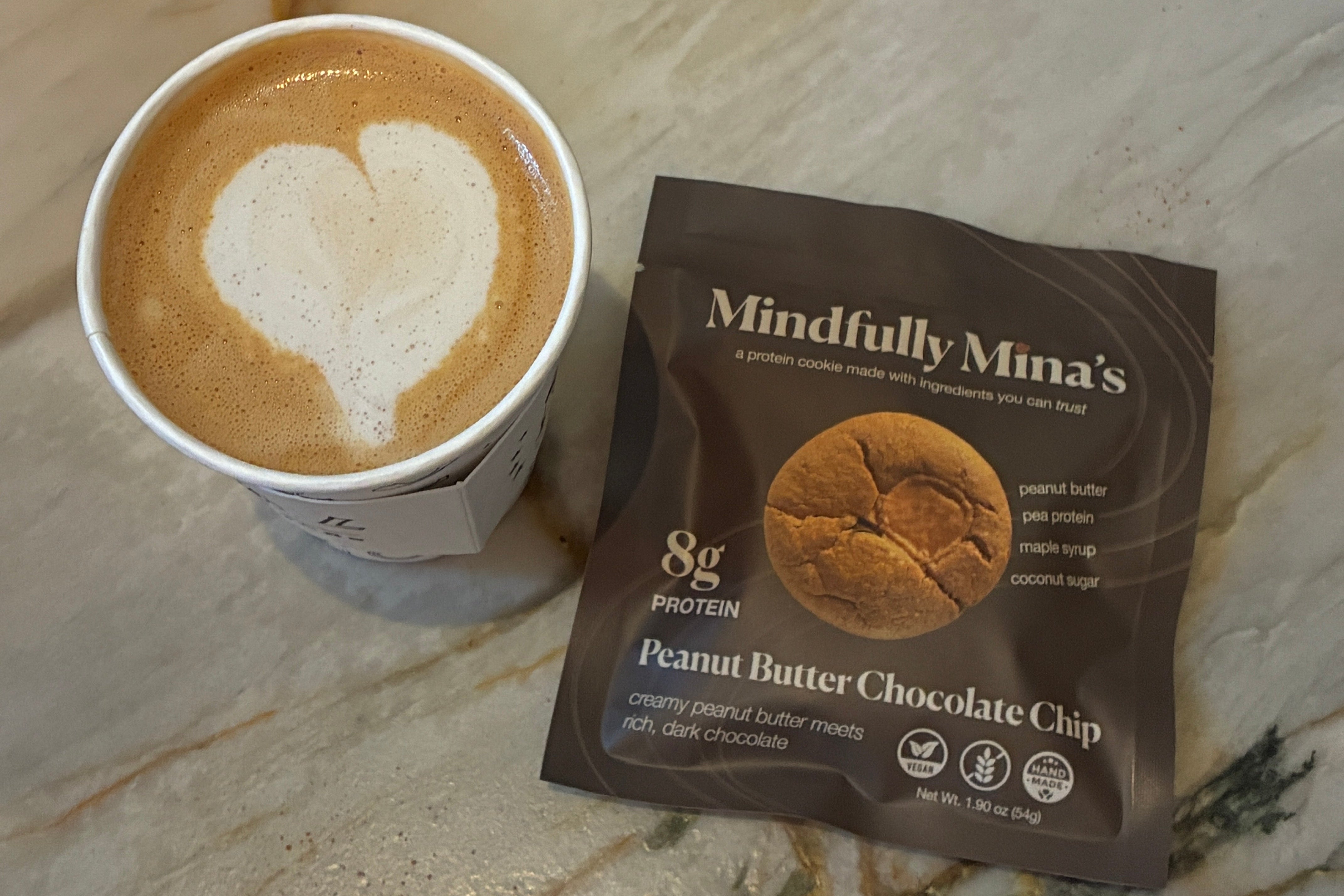 A cup of latte with heart-shaped foam art sits next to a single-serve packet of Mindfully Mina's Peanut Butter Chocolate Chip protein cookie on a polished marble surface.