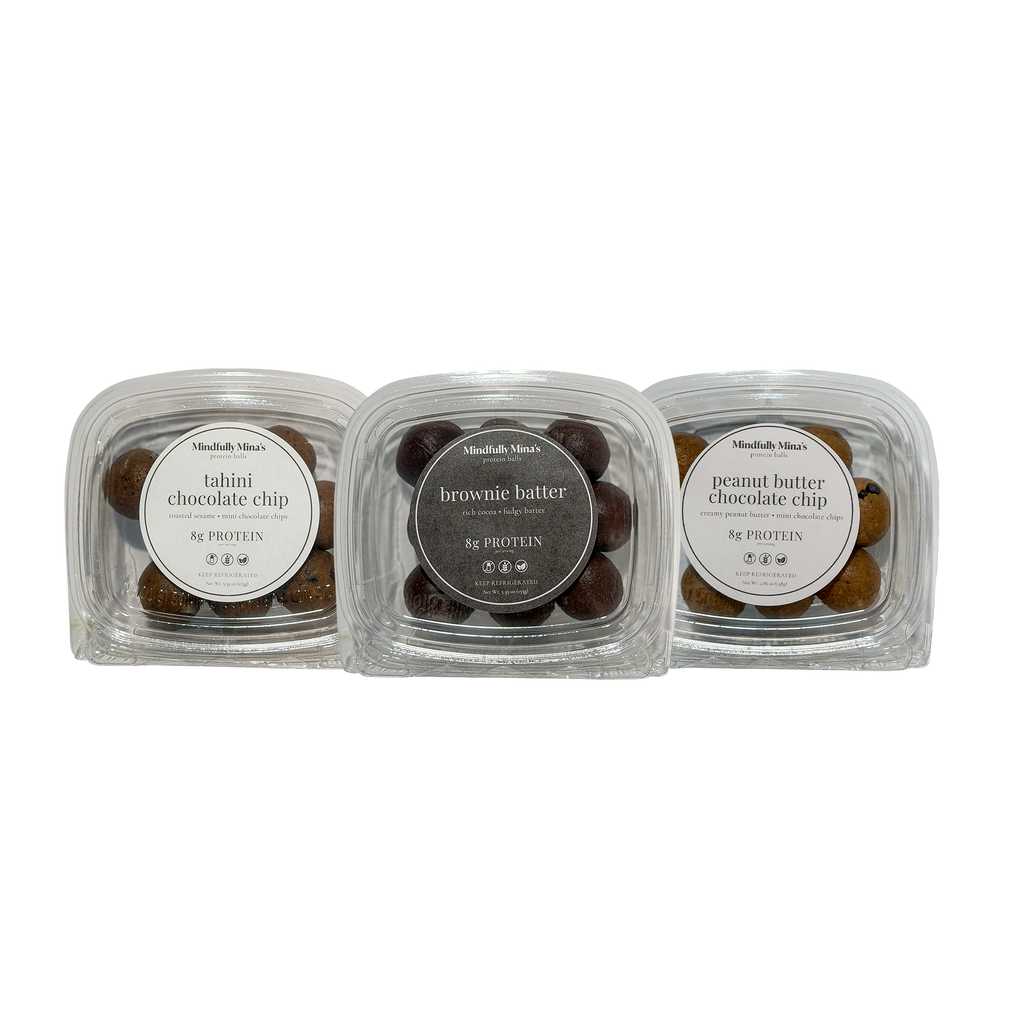 Three clear plastic containers of protein balls in a row, each with a round front label reading “8g protein”: tahini chocolate chip on the left, brownie batter in the middle, and peanut butter chocolate chip on the right. The balls are visible through the clear lids.