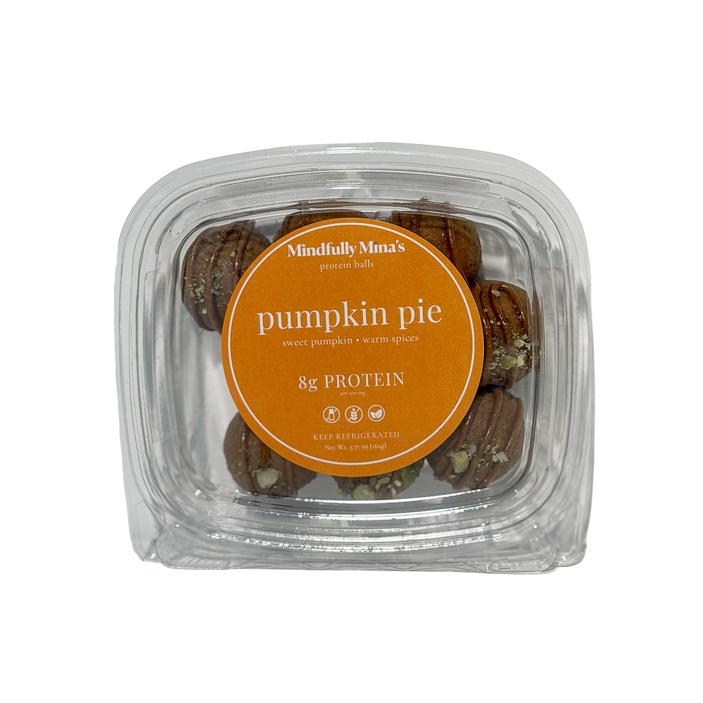 A clear plastic container of pumpkin pie protein balls with a round orange label reading “Mindfully Mina’s protein balls, pumpkin pie, sweet pumpkin, warm spices, 8g protein, keep refrigerated.” The balls are glazed with brown drizzle lines and topped with pumpkin seeds.