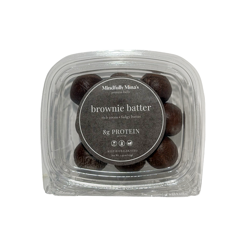 A clear plastic container of dark brownie batter protein balls with a round charcoal-brown label reading “Mindfully Mina’s protein balls, brownie batter, rich cocoa, fudgy batter, 8g protein, keep refrigerated.”