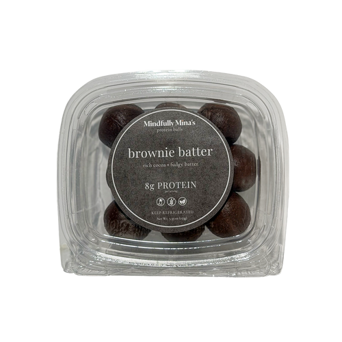 A clear plastic container of dark brownie batter protein balls with a round charcoal-brown label reading “Mindfully Mina’s protein balls, brownie batter, rich cocoa, fudgy batter, 8g protein, keep refrigerated.”