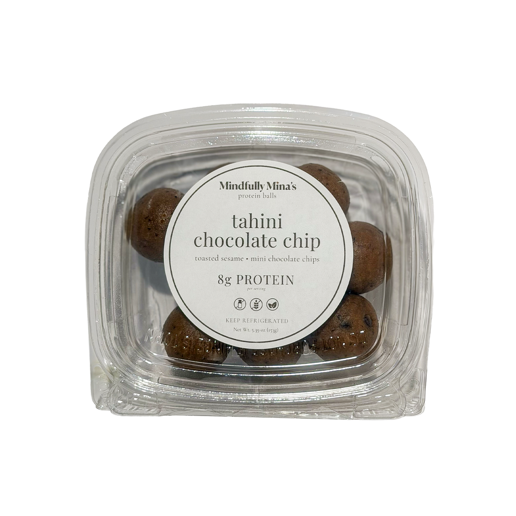 A clear plastic container of tahini chocolate chip protein balls with a round white label reading “Mindfully Mina’s protein balls, tahini chocolate chip, toasted sesame, mini chocolate chips, 8g protein, keep refrigerated.”